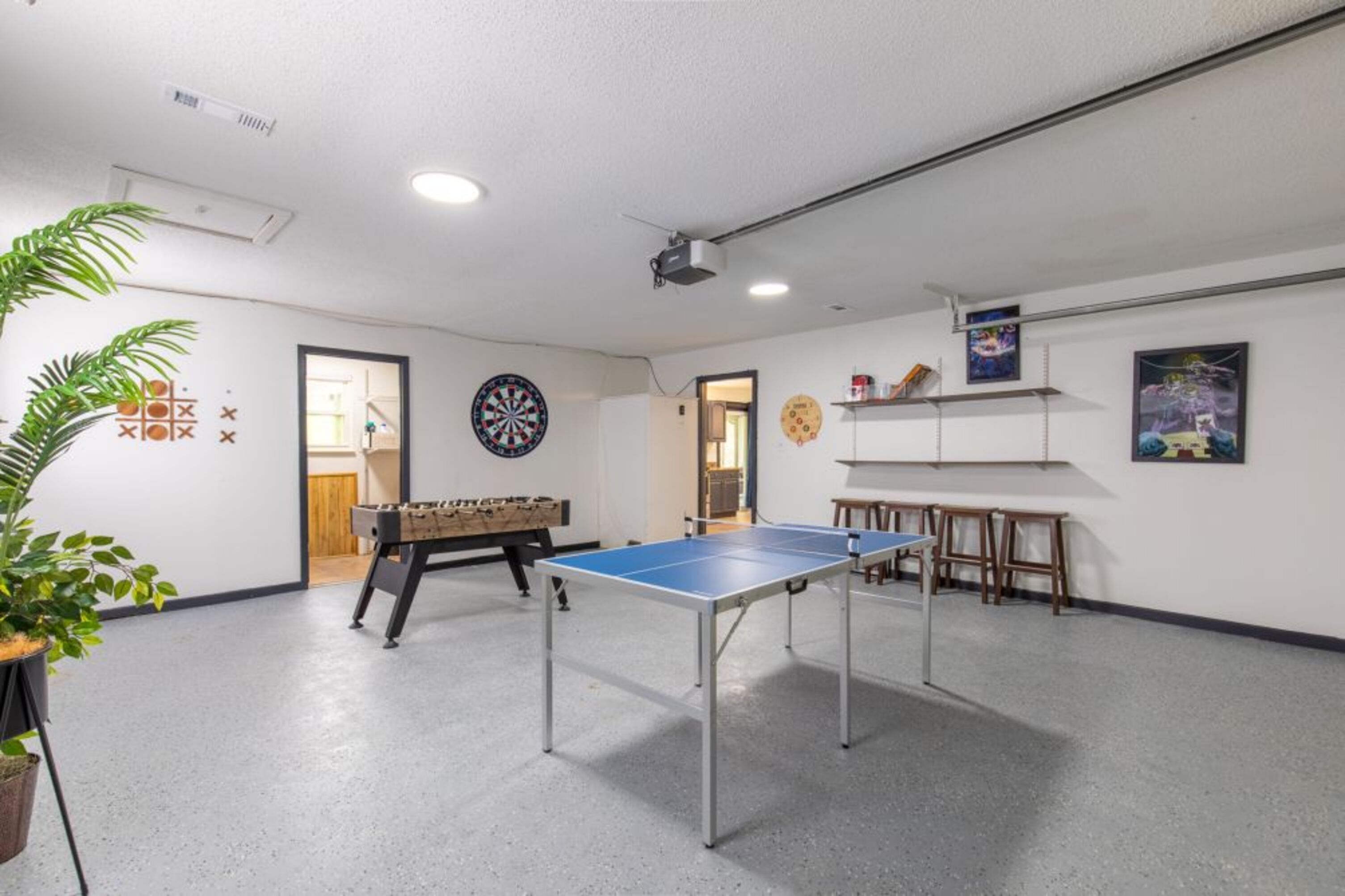 A room with a foosball table, ping pong table, dartboard, and shelves with items.