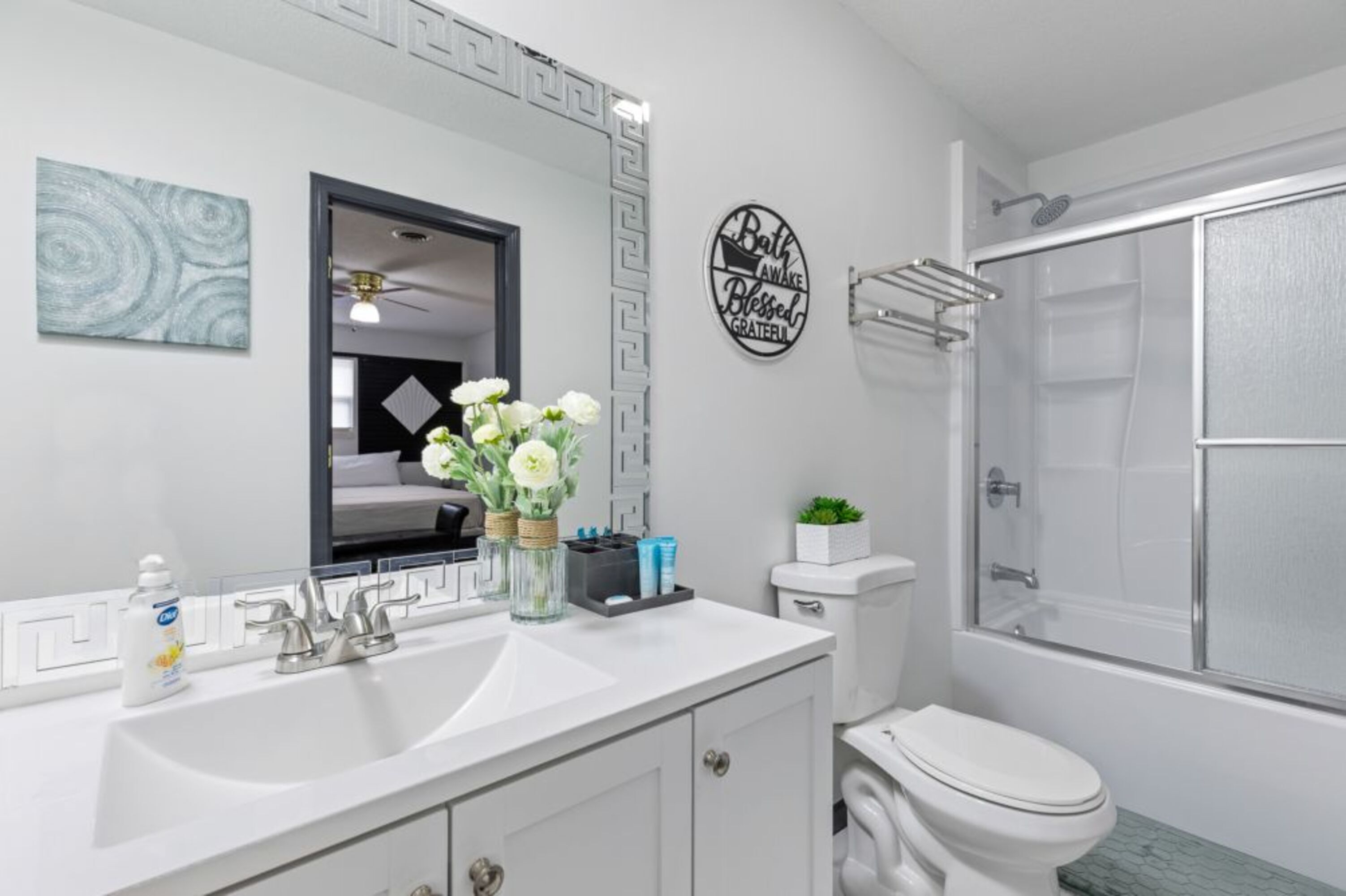 A bathroom with a white sink, mirror, toilet, and shower. There is a decorative sign on the wall and a vase with flowers on the counter.