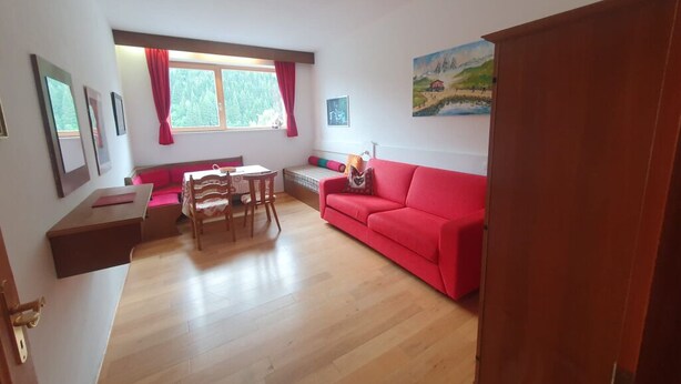 Ein Wohnzimmer mit rotem Sofa, Holzboden, einem Esstisch mit Stühlen und einem Fenster mit roten Vorhängen.