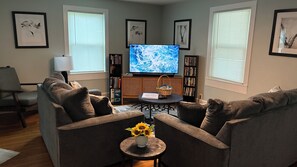 Een woonkamer met een bank, een bijzettafeltje met een vaas zonnebloemen, een televisie en een boekenkast.