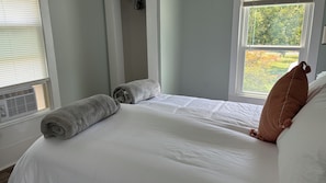 Cama bem arrumada com lençóis brancos e duas toalhas enroladas, uma janela com persianas e vista para o verde lá fora.