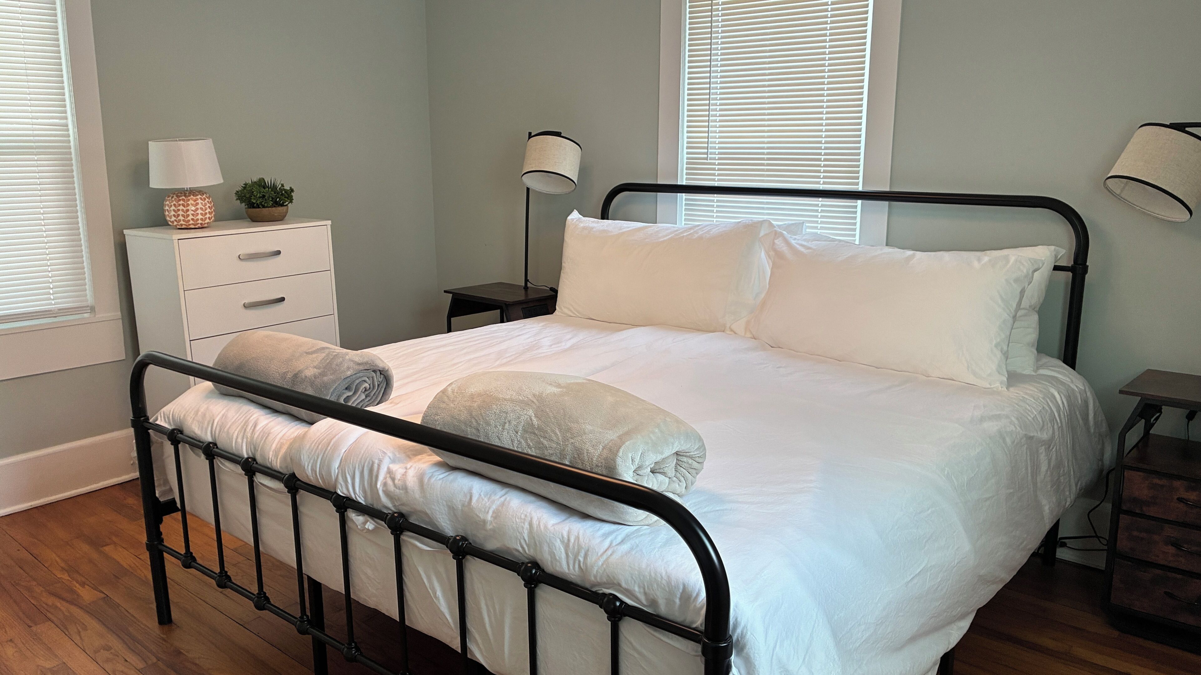 Uma cama bem arrumada com roupa de cama branca e duas almofadas, uma mesa de cabeceira com abajur e uma pequena planta em vaso.