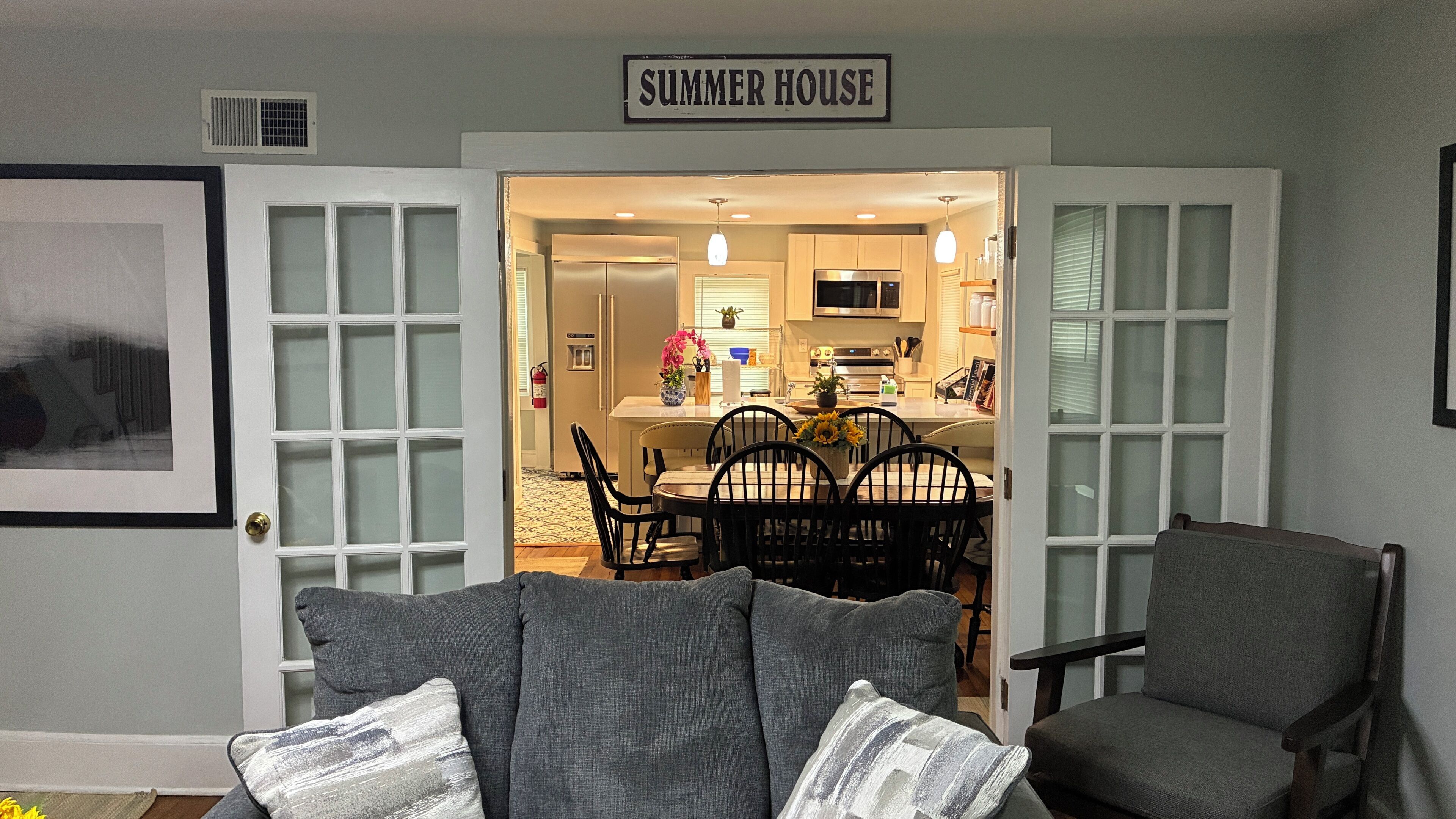 Una sala de estar con comedor, un letrero que dice 'SUMMER HOUSE', y una cocina visible a través de una puerta abierta.