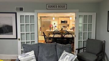 Una sala de estar con comedor, un letrero que dice 'SUMMER HOUSE', y una cocina visible a través de una puerta abierta.