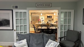 Uma sala de estar com área de jantar, uma placa com a inscrição 'SUMMER HOUSE' e cozinha visível por uma porta aberta.