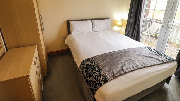 A hotel room with a bed, bedside table, lamp, and a view through a window. - Holiday Cottage 1 (Skegness)