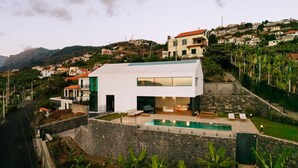 Una casa moderna de dos pisos con piscina, rodeada por un muro de piedra y abundante vegetación.