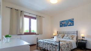Une chambre à coucher avec un lit, des tables de chevet, une fenêtre avec des rideaux, un radiateur, un bureau avec une plante et un tableau au mur.