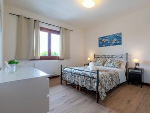 Ein Schlafzimmer mit Bett, Nachttischen, einem Fenster mit Vorhängen, einem Heizkörper, einem Schreibtisch mit Pflanze und einem Bild an der Wand.