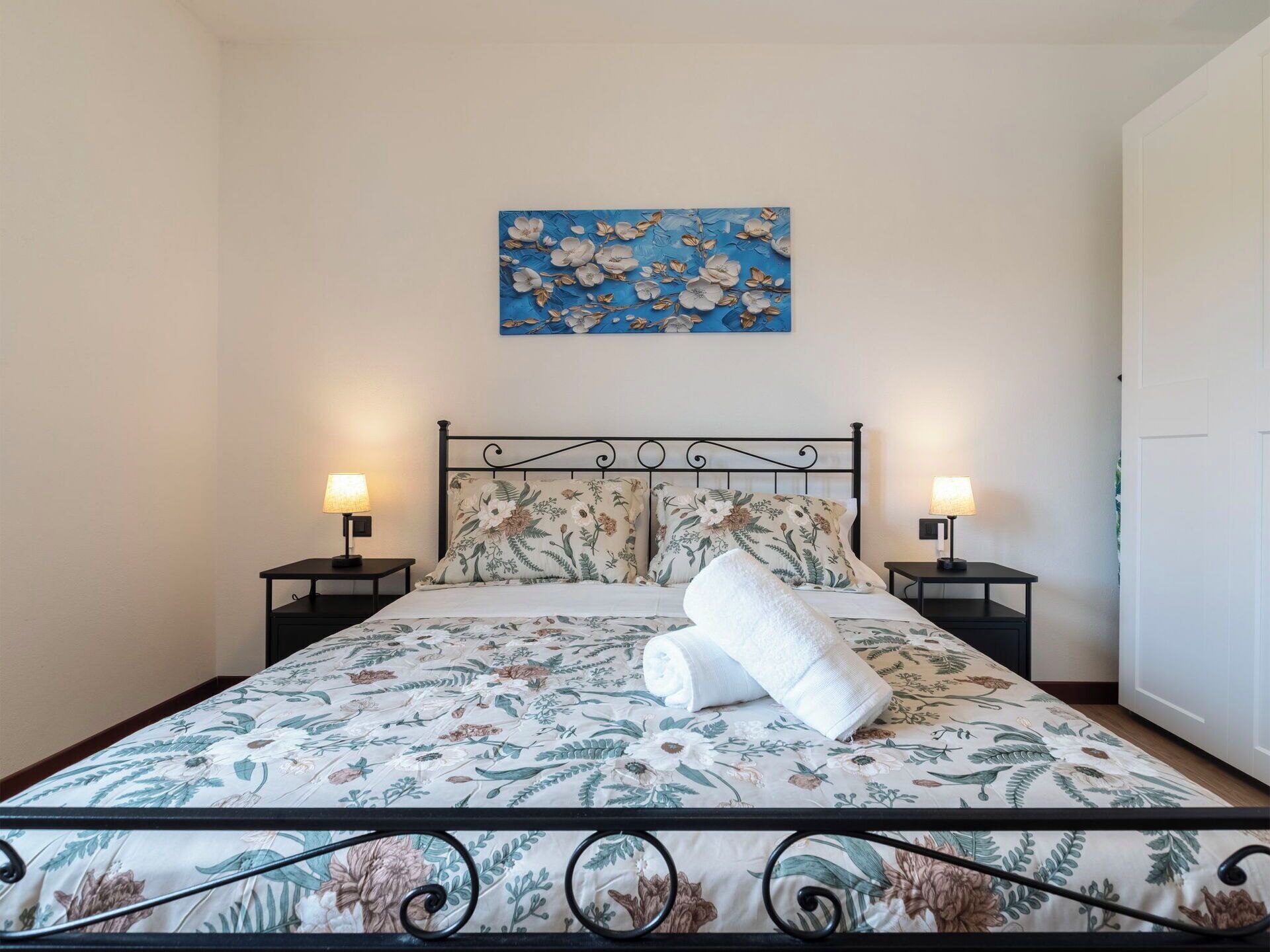A neatly made bed with floral bedding, two bedside lamps, and a blue floral painting above.