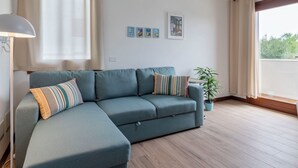Un salon avec un canapé d’angle, une lampe, une plante en pot et une fenêtre avec des rideaux.