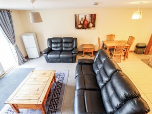 A living room with a black leather sofa, a wooden coffee table, a dining table with chairs, a white refrigerator, and a painting on the wall. - Holiday Cottage 3 (Skegness)