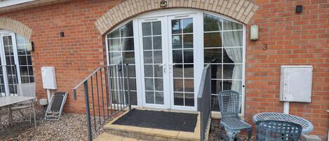 Un edificio in mattoni con un ingresso ad una porta in vetro, una piccola area esterna con due sedie e una panchina in pietra.