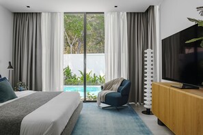 Quarto moderno com cama, televisão, poltrona e vista para uma piscina.