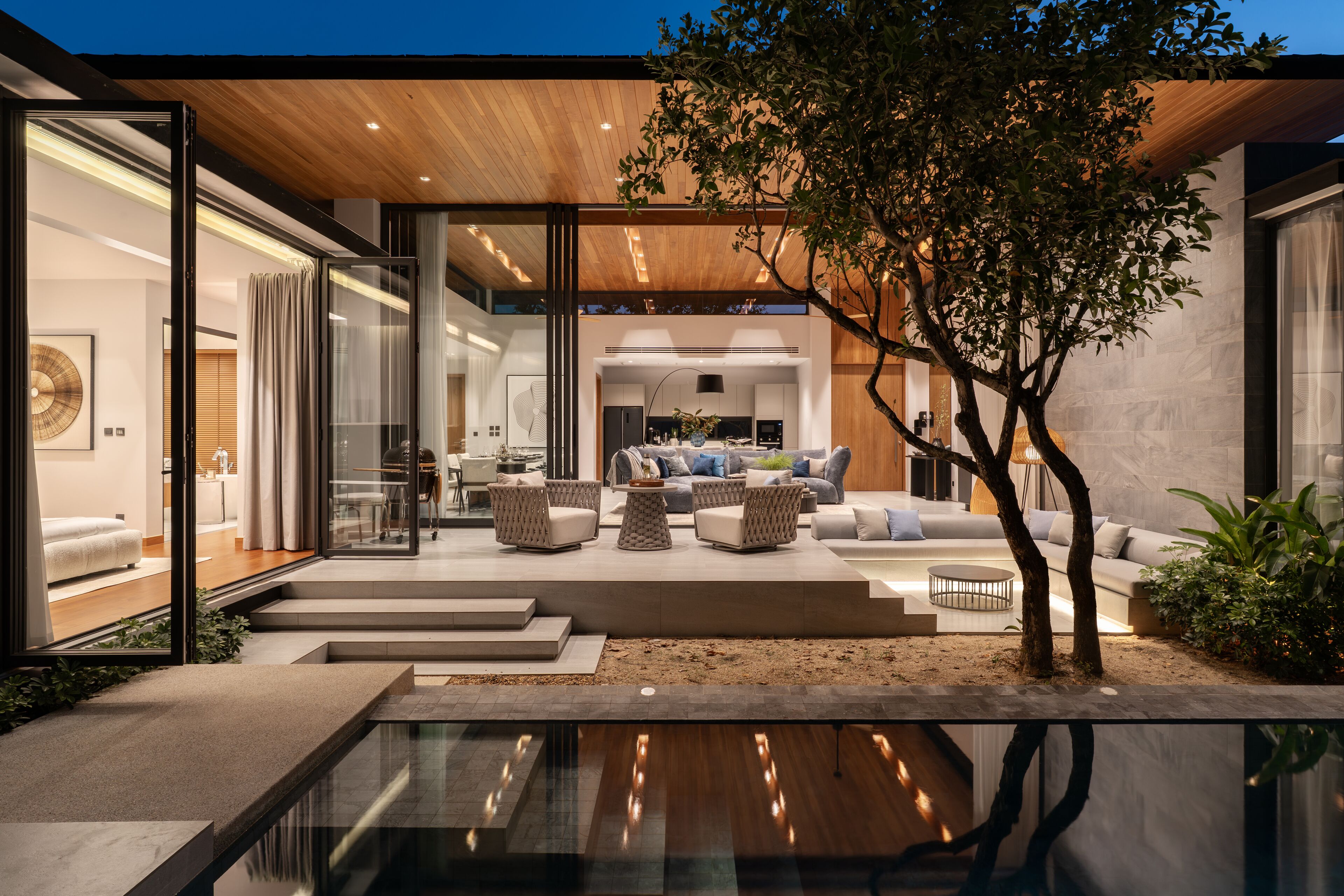 A modern house with a glass entrance, a dining area with sofas, and a tree in the center.