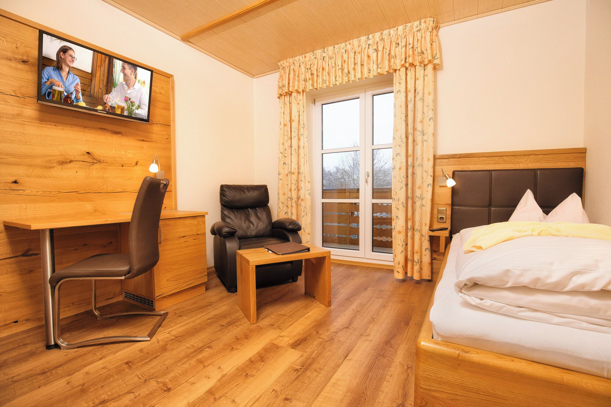 A hotel room with a wooden desk, a massage chair, a bed, and a large window with curtains.