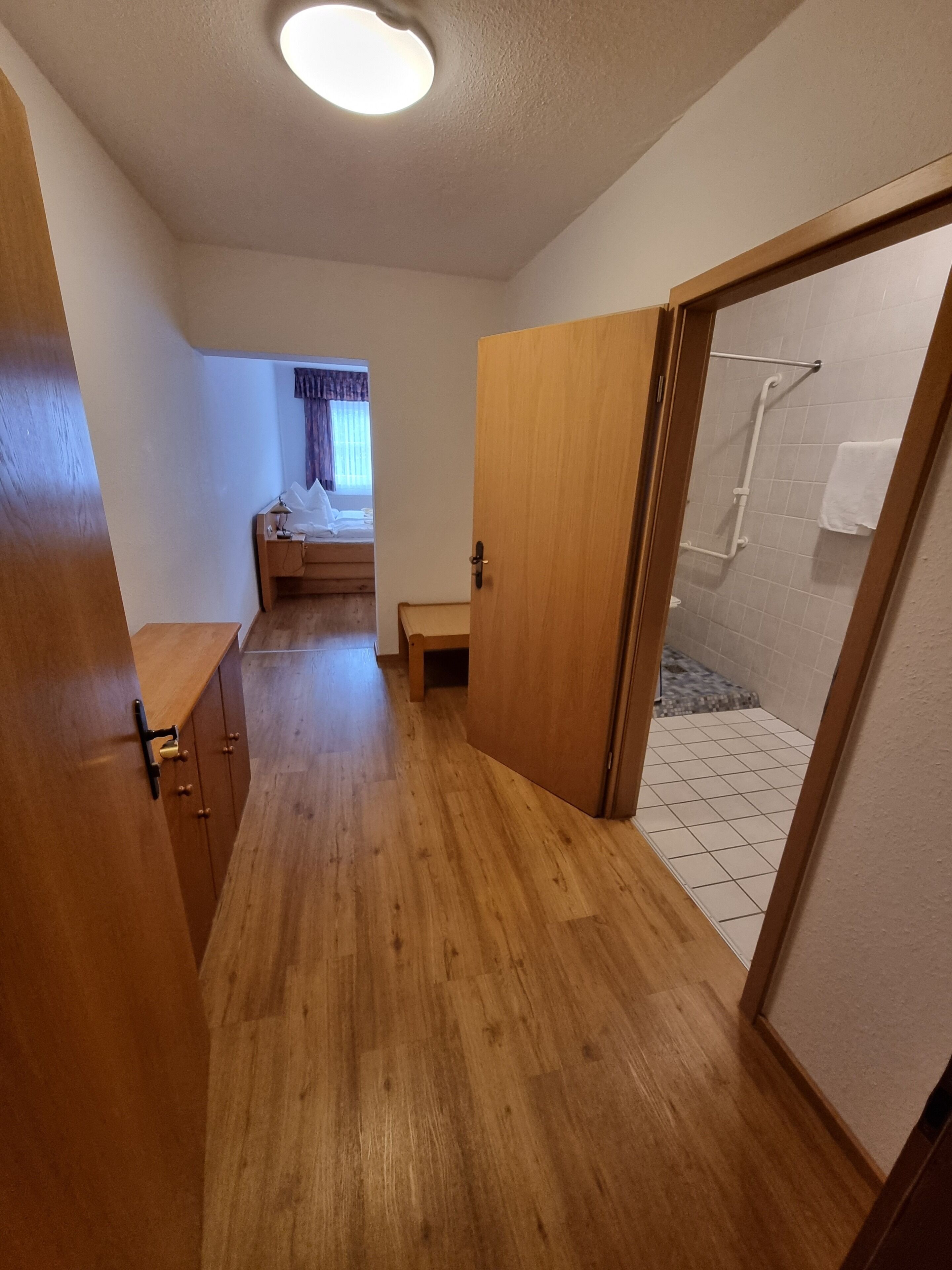Ein schmaler Flur mit Holzboden führt zu einem Schlafzimmer mit Bett und einem Fenster mit Vorhängen.