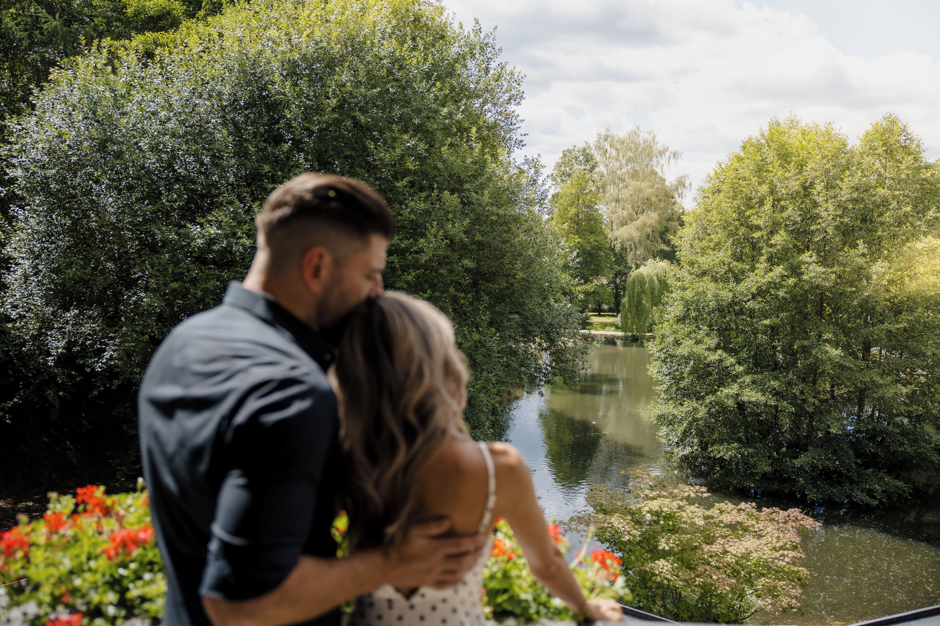 A couple embracing by a riverside with lush greenery and a calm water body.