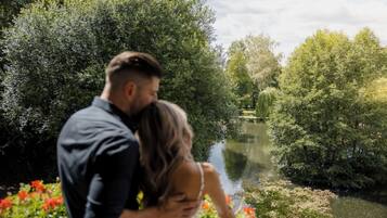 A couple embracing by a riverside with lush greenery and a calm water body.