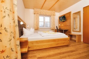 A bedroom with a wooden bed, a bedside table, a TV mounted on the wall, and a window with curtains. - Der Glaserhof (Lam)