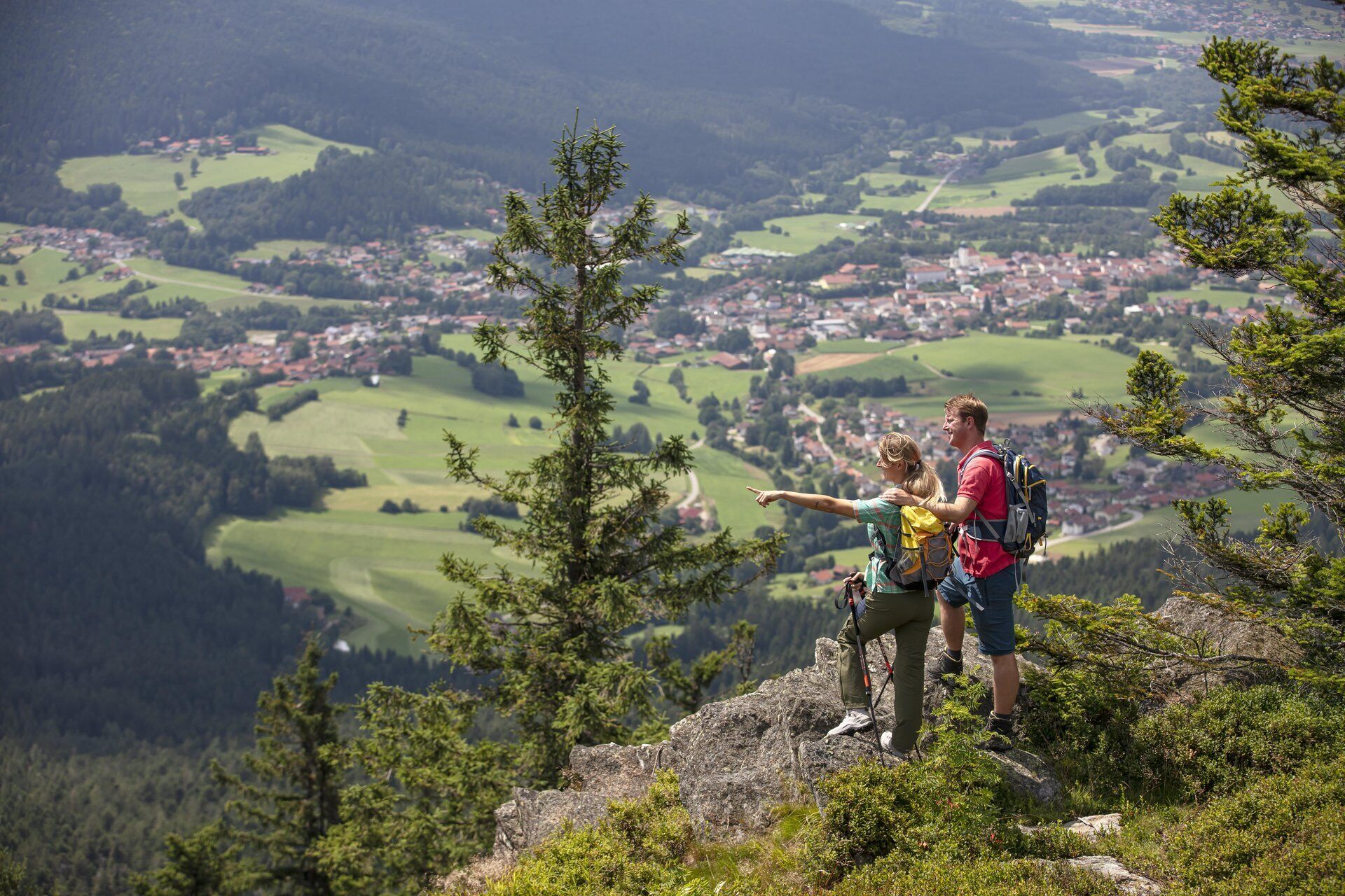 A couple standing on a mountain peak overlooking a valley with a town below.