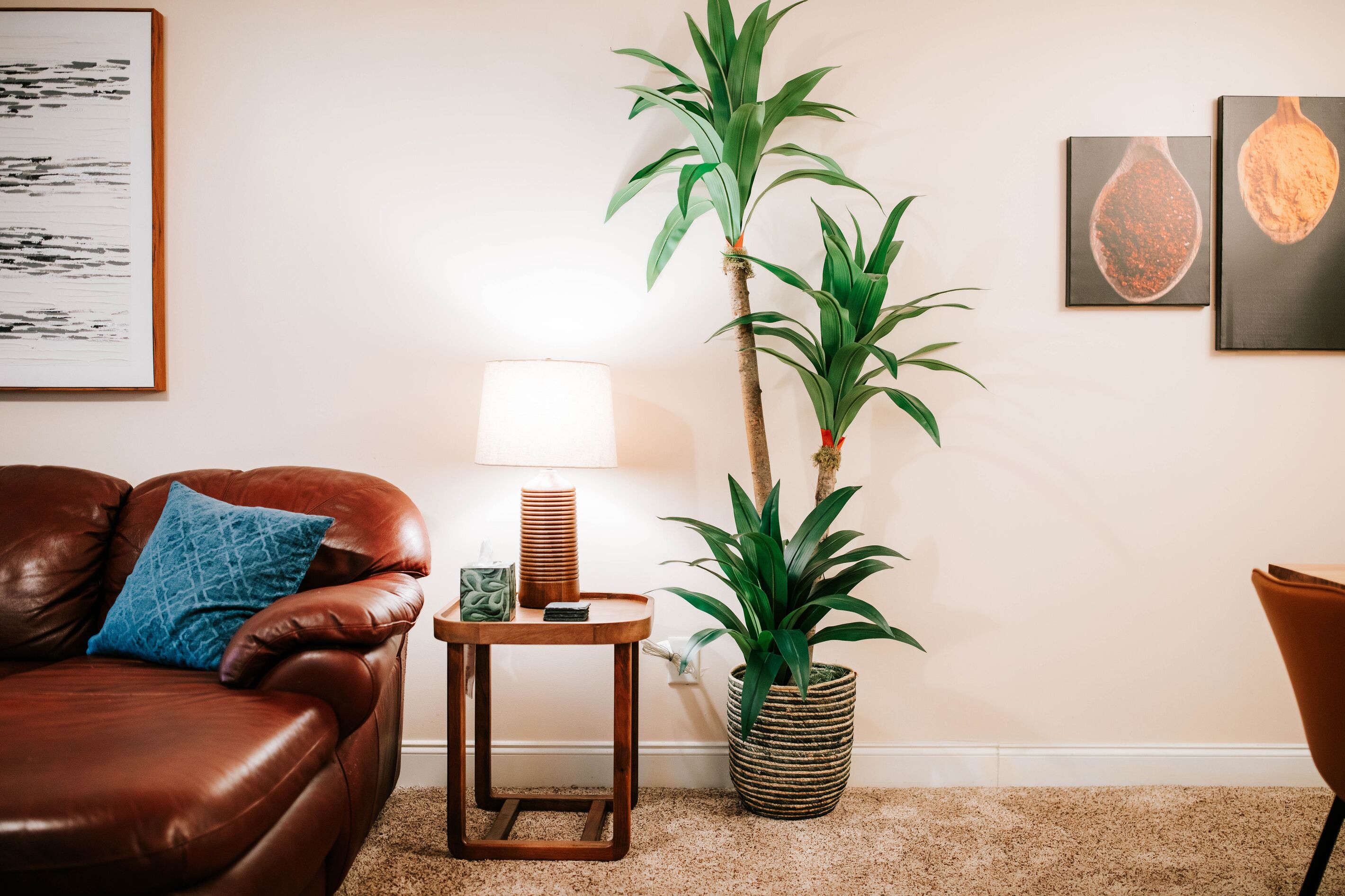 A living room with a brown leather sofa, a small wooden side table, a tall potted plant, a wall-mounted lamp, and framed artwork.