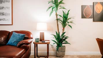 A living room with a brown leather sofa, a small wooden side table, a tall potted plant, a wall-mounted lamp, and framed artwork.