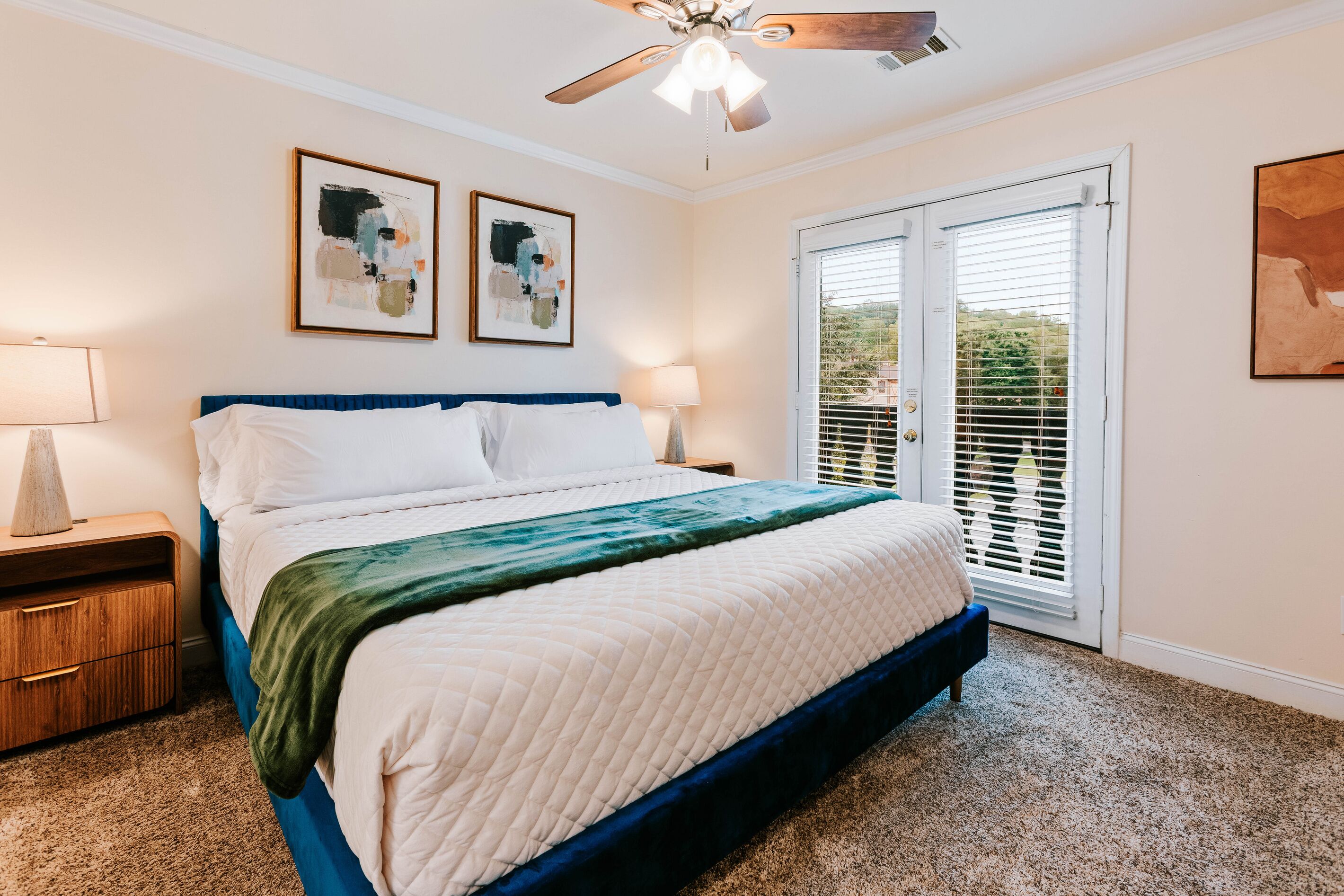 A bedroom with a bed, a nightstand, a lamp, two abstract paintings, a ceiling fan, and a sliding glass door.