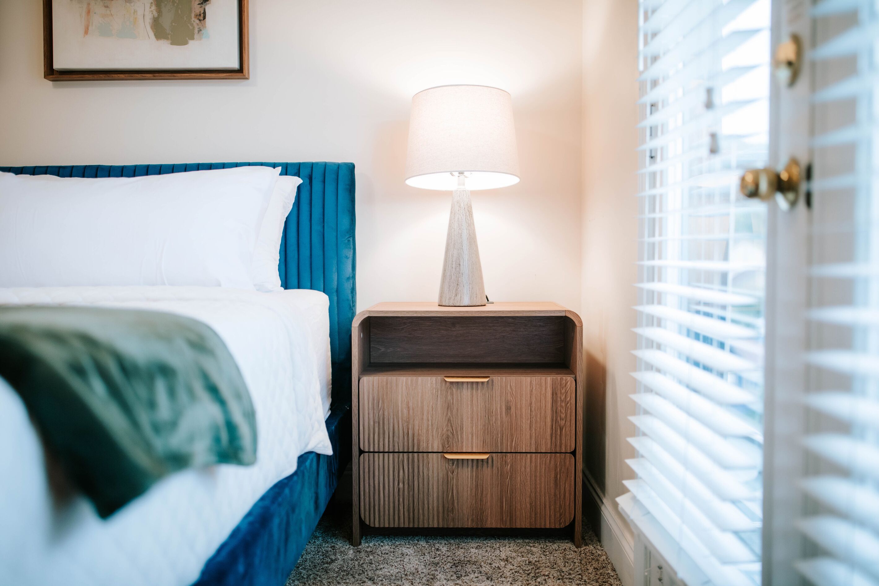 A neatly made bed with a blue headboard, a bedside table with a lamp, and a window with blinds.