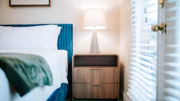 A neatly made bed with a blue headboard, a bedside table with a lamp, and a window with blinds.
