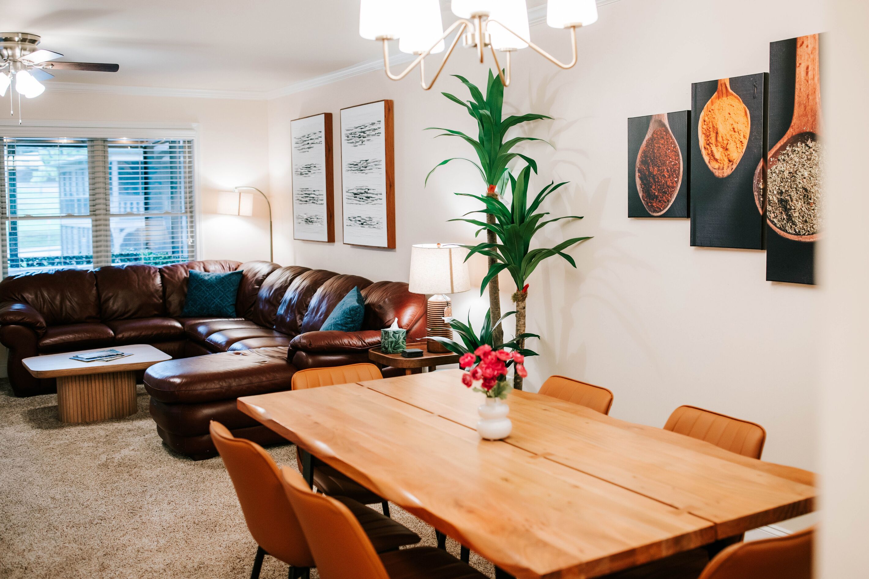 A modern living room with a brown leather sofa, a wooden dining table with chairs, a potted plant, and artwork on the walls.