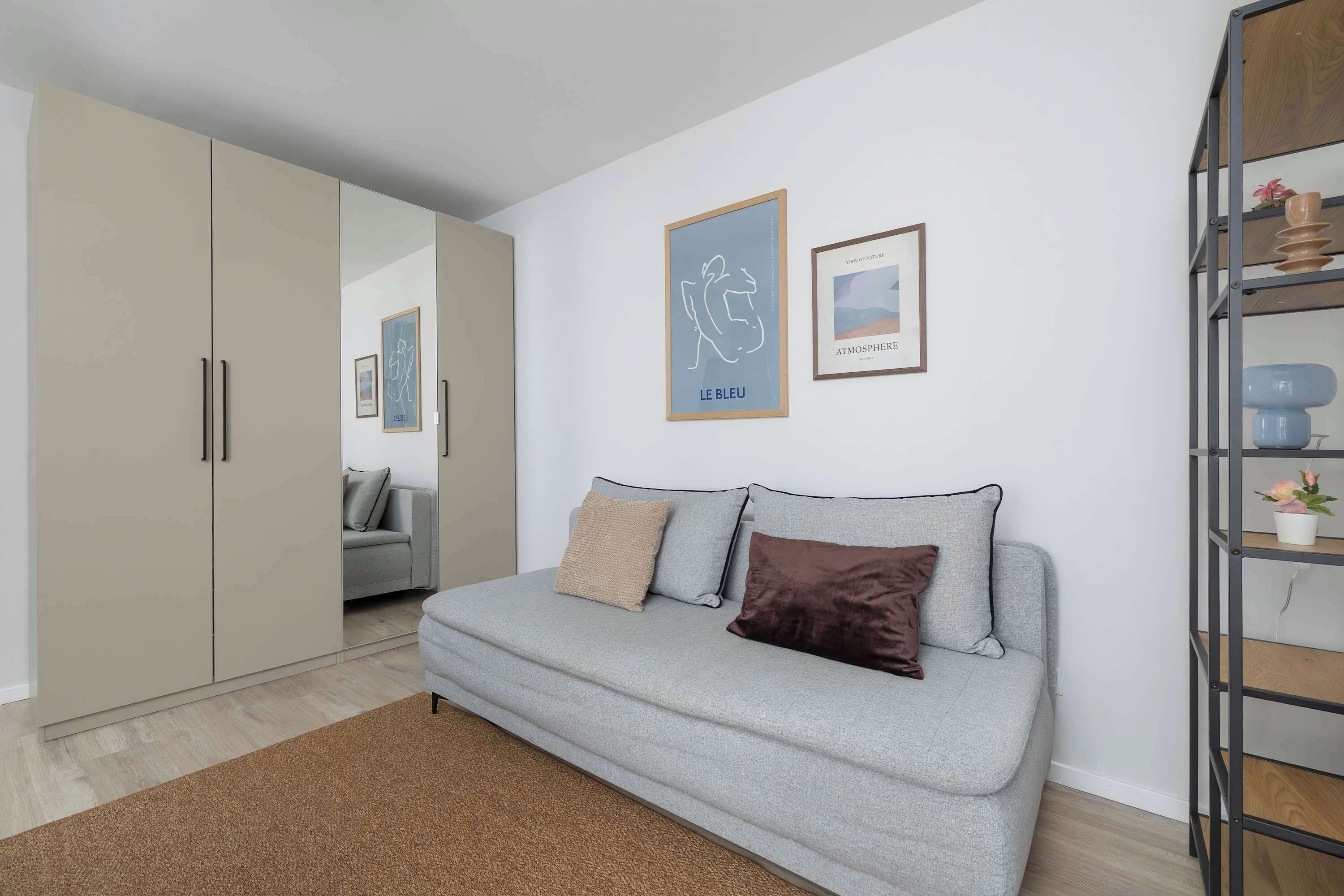 Una sala de estar moderna con un sofá gris, dos almohadas, un tapete marrón y piso de madera. Hay obras de arte enmarcadas en la pared y un armario con espejo.