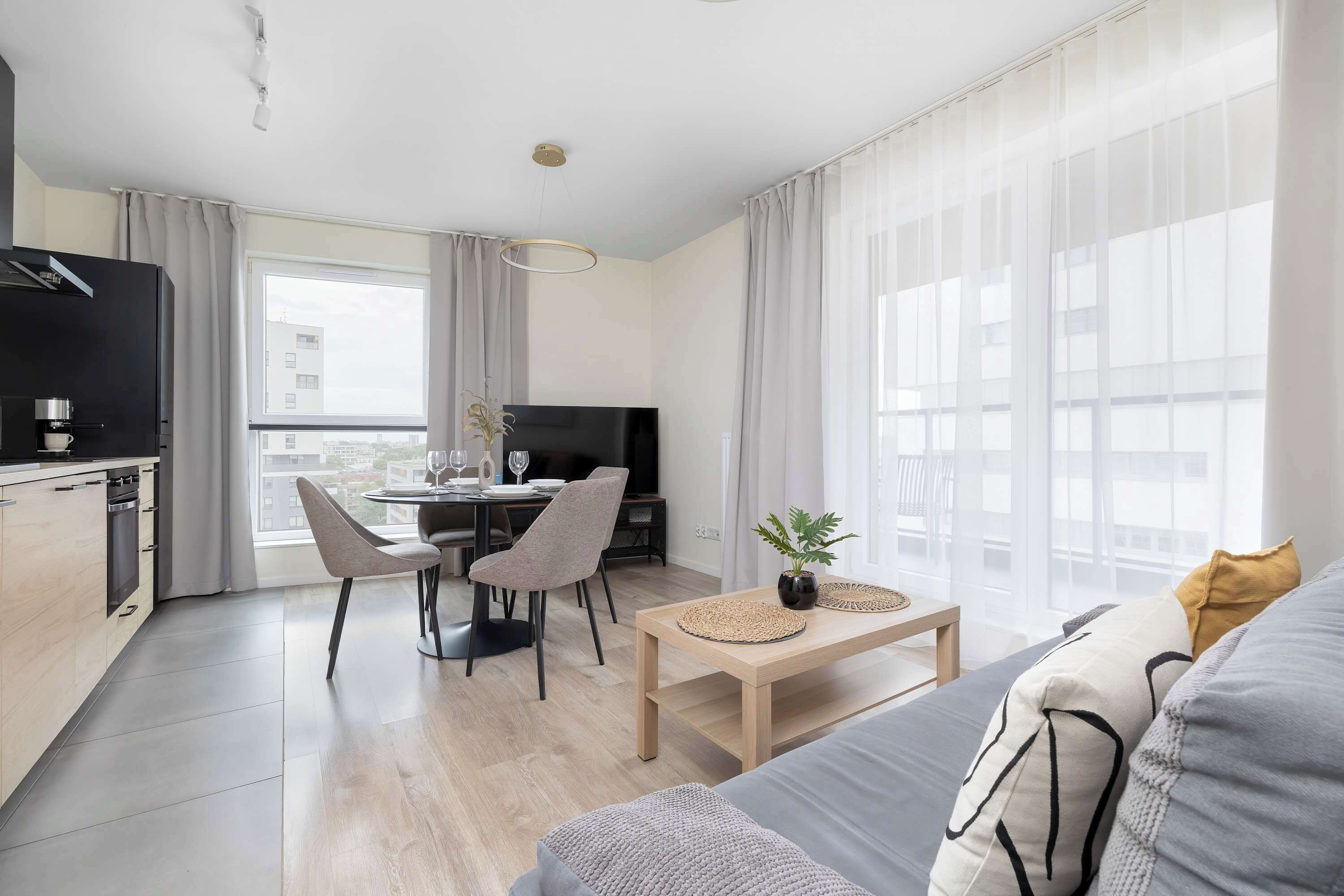Ett modernt vardagsrum med en soffa, en matplats med stolar, ett kök med en kaffemaskin och stora fönster.