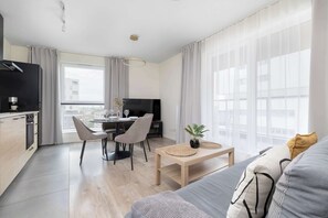 Een moderne woonkamer met een bank, een eethoek met stoelen, een keuken met een koffiemachine, en grote ramen.