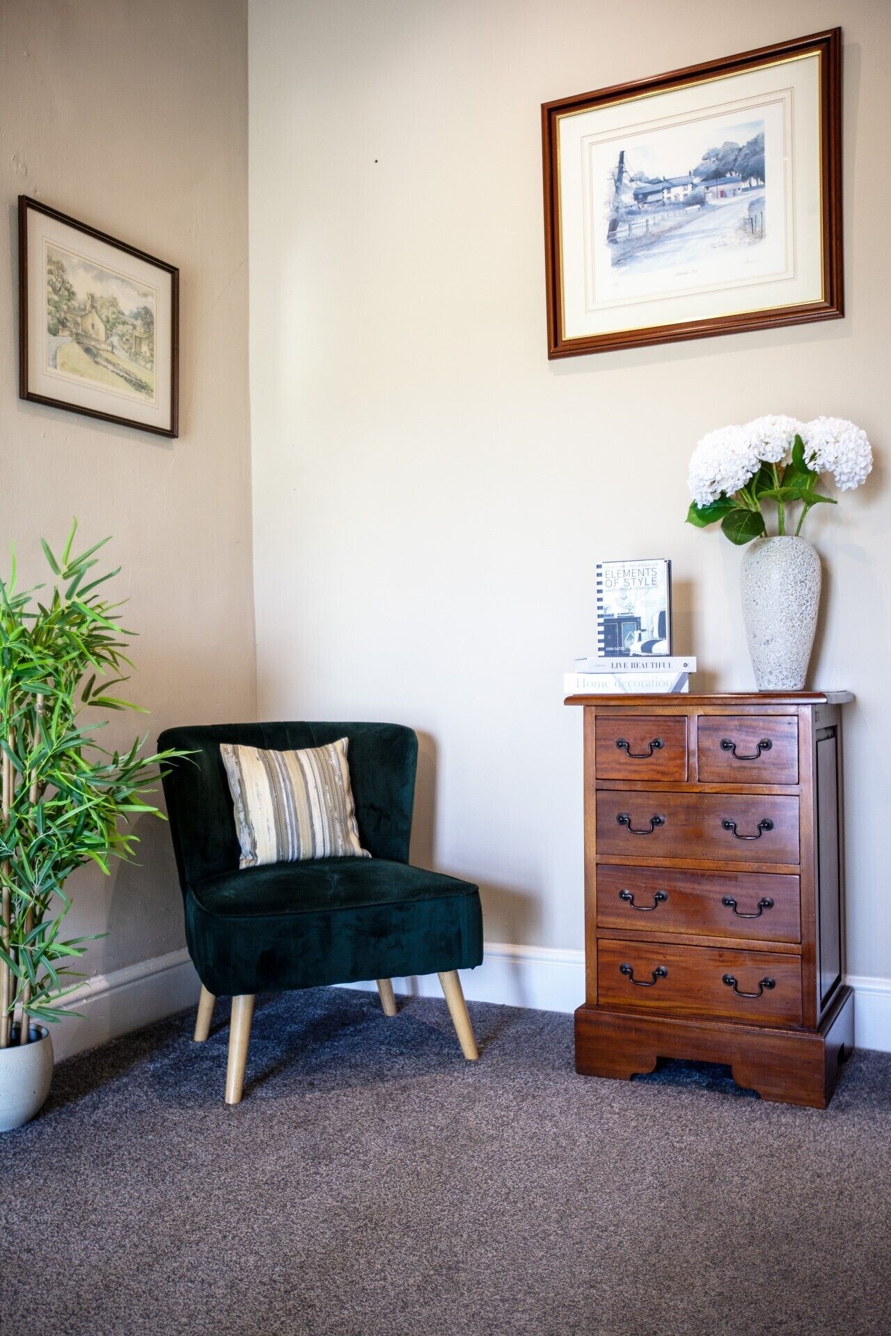 A room with a dark green velvet armchair, a wooden dresser with multiple drawers, a potted bamboo plant, and two framed artworks on the wall.