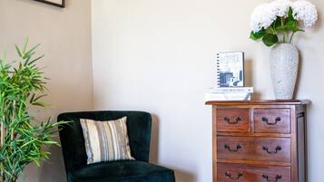 A room with a dark green velvet armchair, a wooden dresser with multiple drawers, a potted bamboo plant, and two framed artworks on the wall.