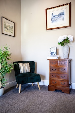 Una habitación con un sillón tapizado en verde oscuro, un aparador de madera con varios cajones, una planta de bambú en maceta y dos cuadros en la pared.
