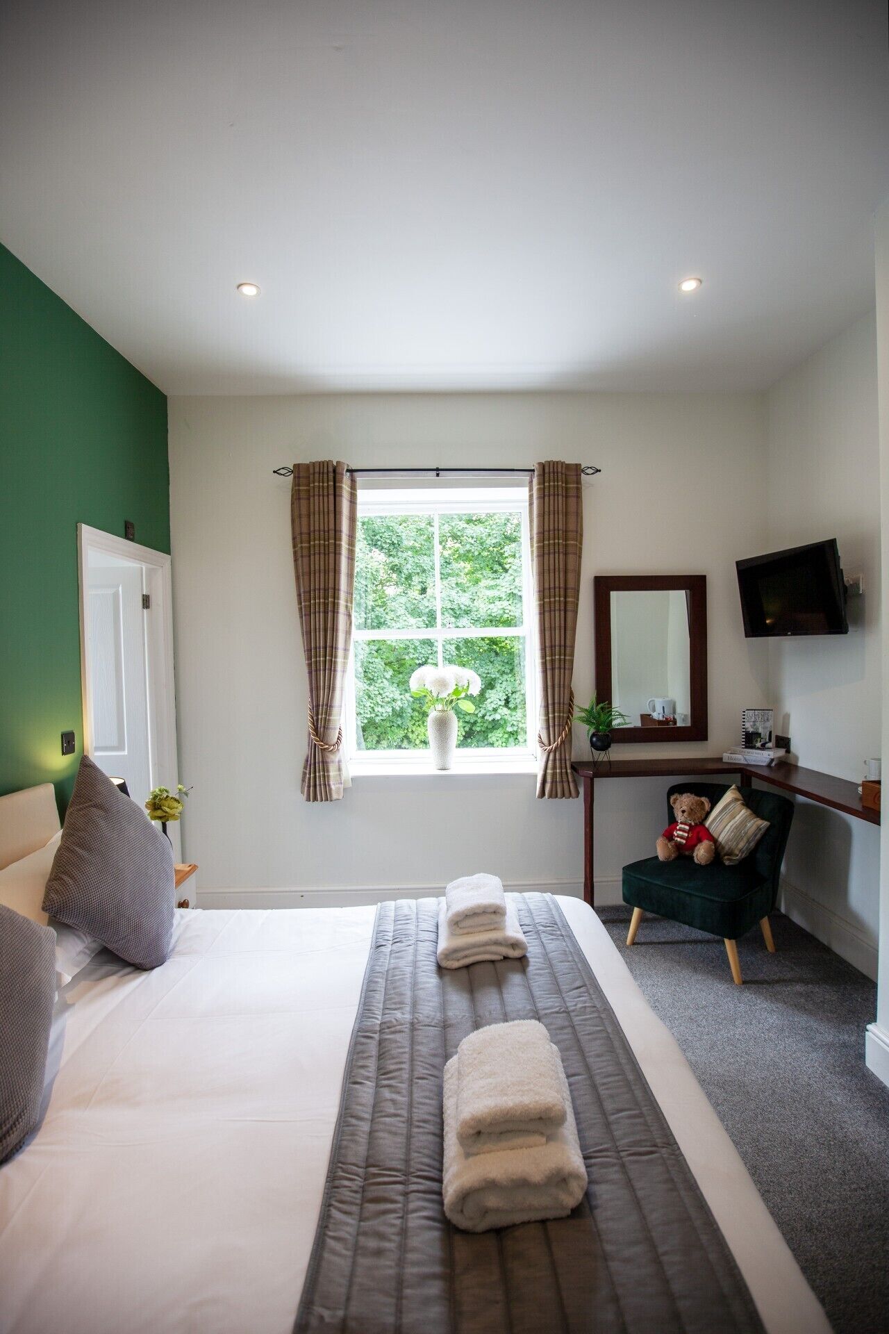 A hotel room with a bed, a desk, a chair, a window with curtains, and a flat-screen TV.