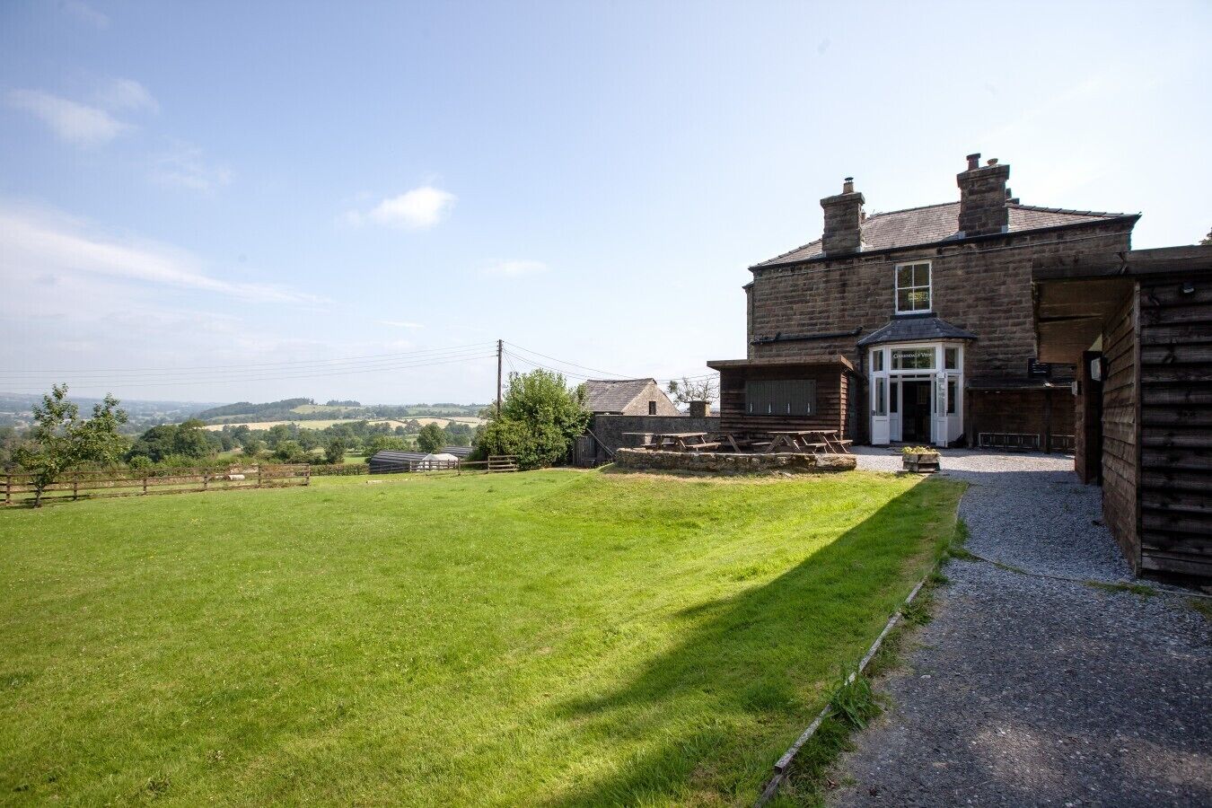 A two-story stone building with a front entrance, a grassy lawn, and a view of the countryside.