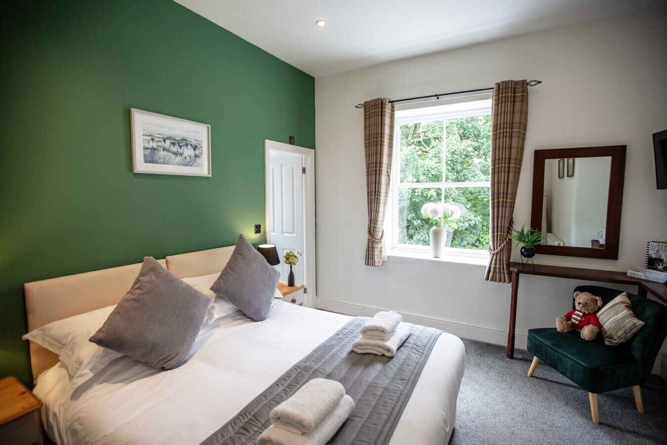 A hotel room with a bed, a bedside table, a chair, a window with curtains, a mirror, and a picture on the wall.