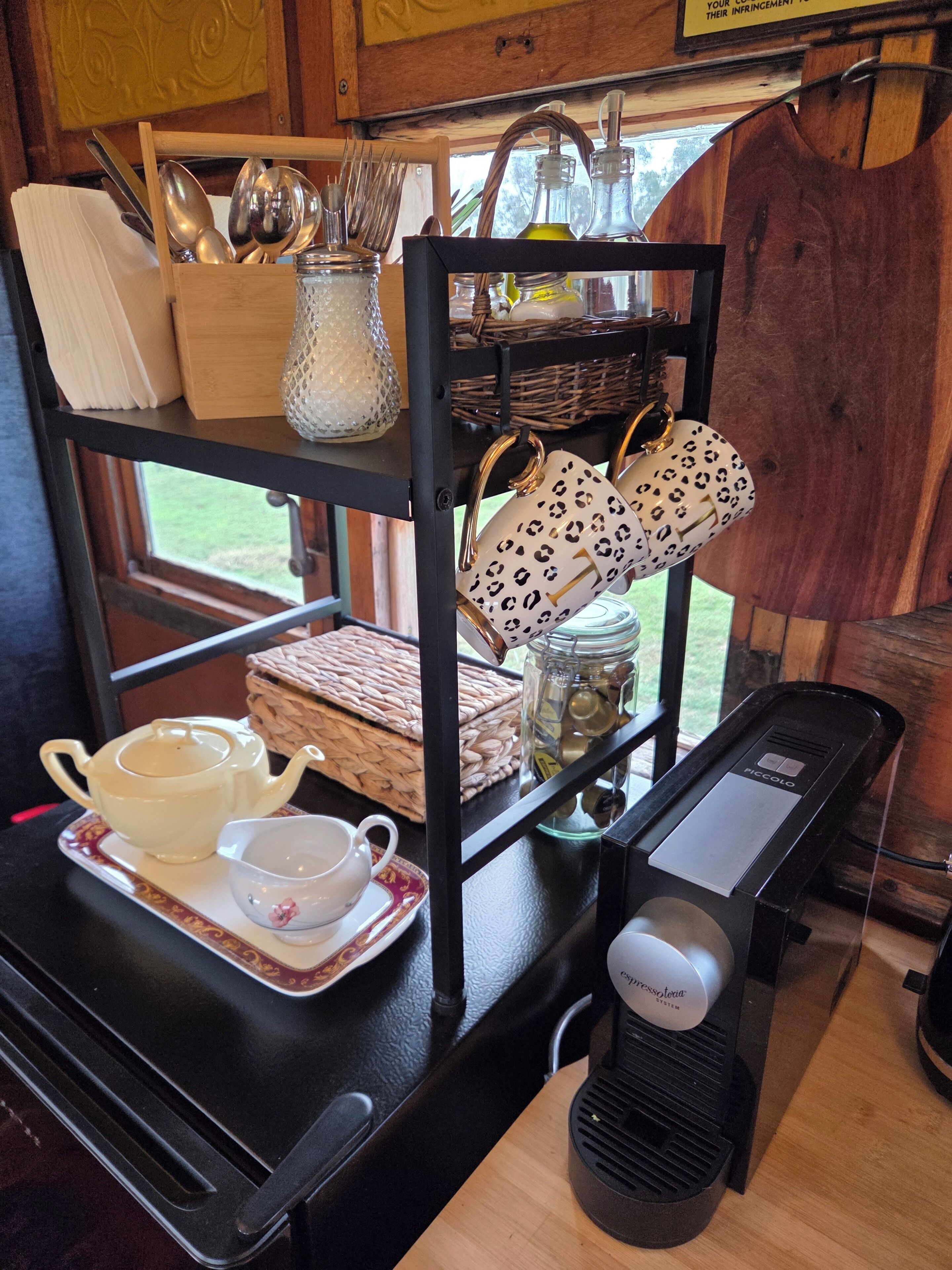 Un área de cocina con cafetera, tetera, tazas y un estante con utensilios y un tarro.