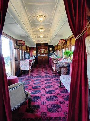A train carriage with a central aisle, tables, chairs, and a patterned carpet.