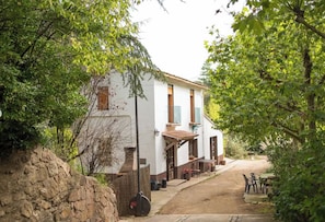 A white building with a stone wall, a wooden door, and a paved path leading to a table and chairs.