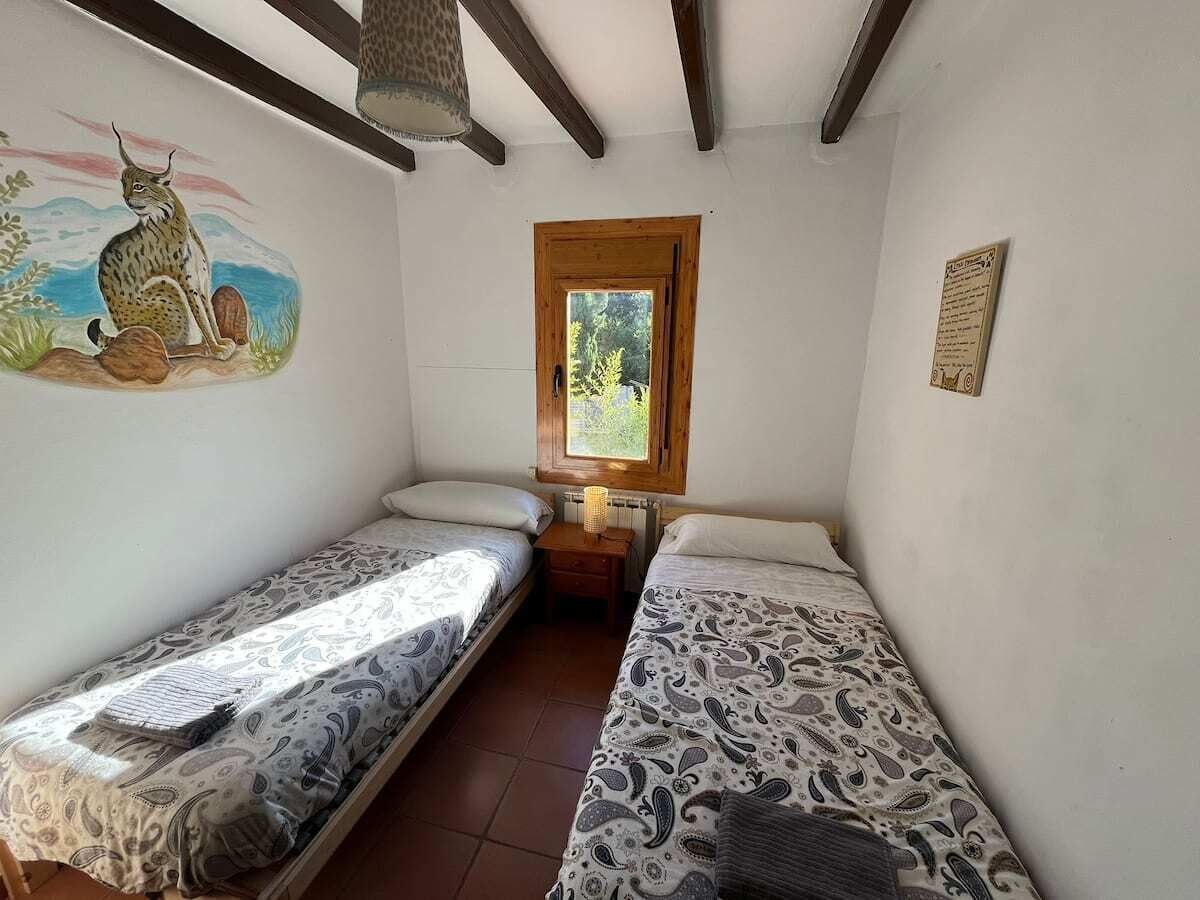 A small room with two single beds, a wooden-framed window, and a wall-mounted lamp.