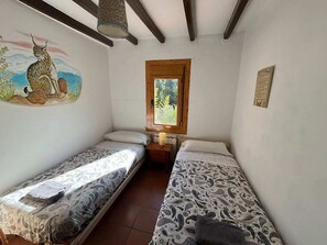 A small room with two single beds, a wooden-framed window, and a wall-mounted lamp.