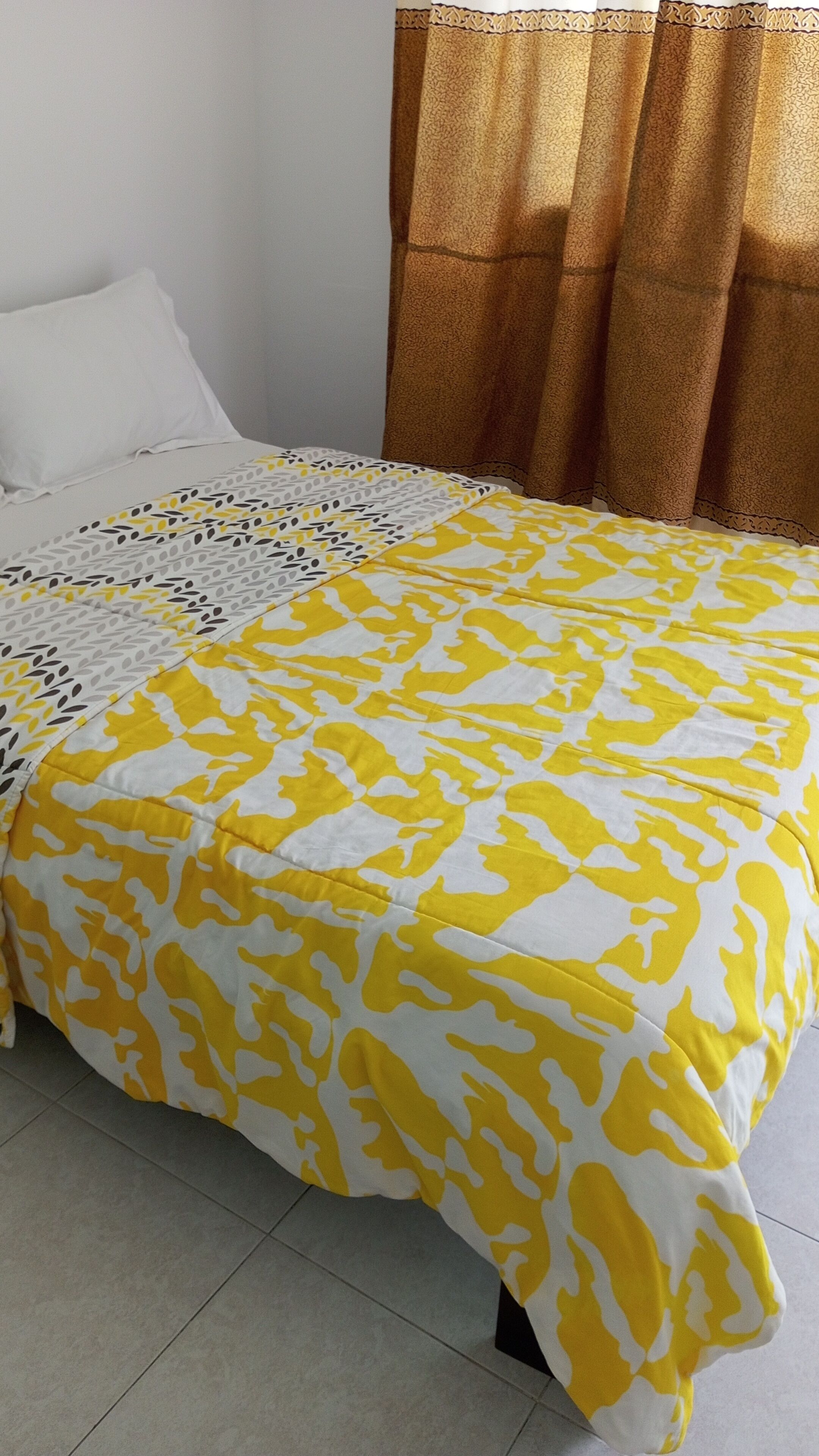 A bed with a patterned bedspread, a white pillow, and a brown curtain.