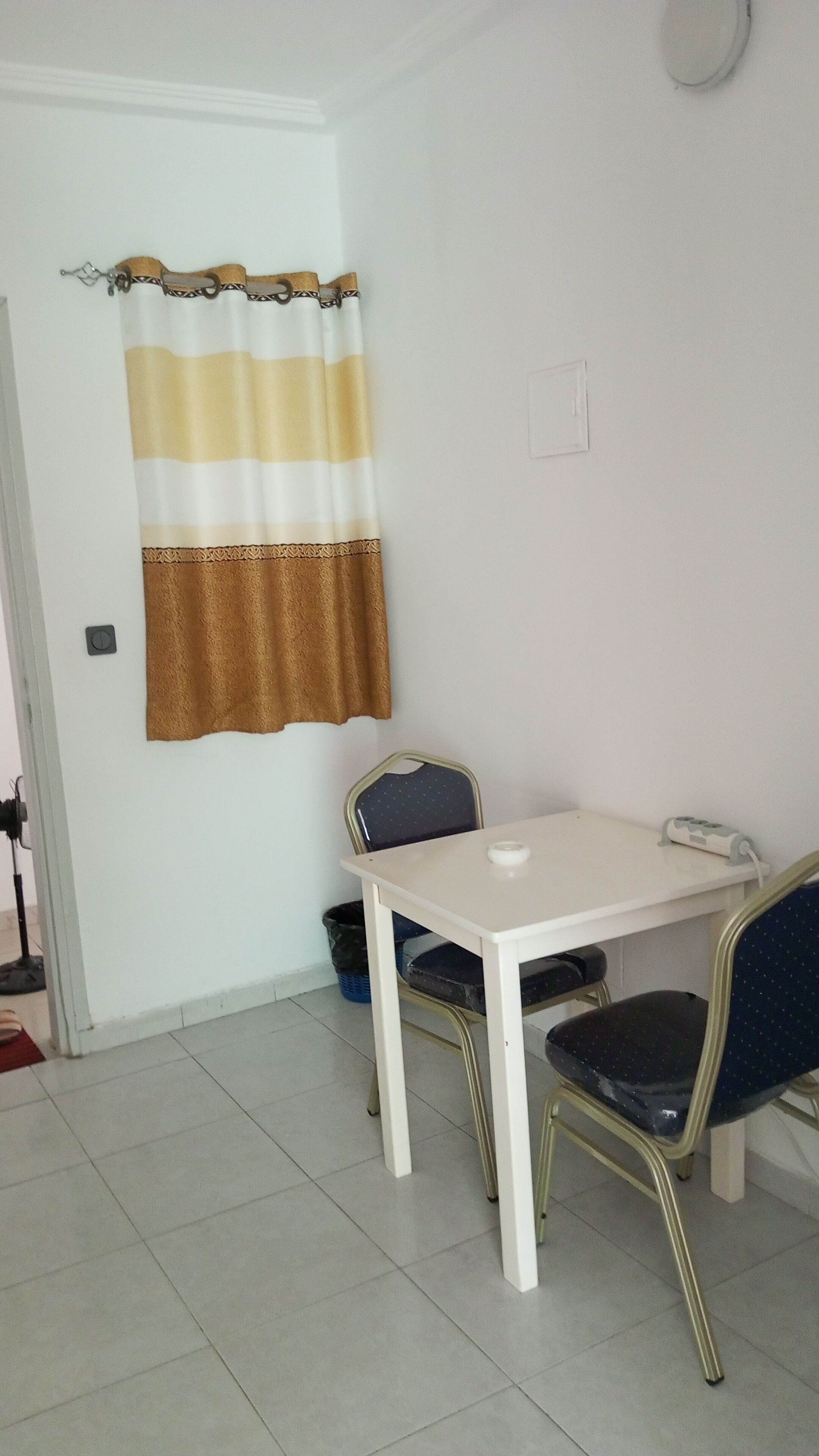 A small dining area with a white table and two chairs, a striped curtain, and a wall socket.