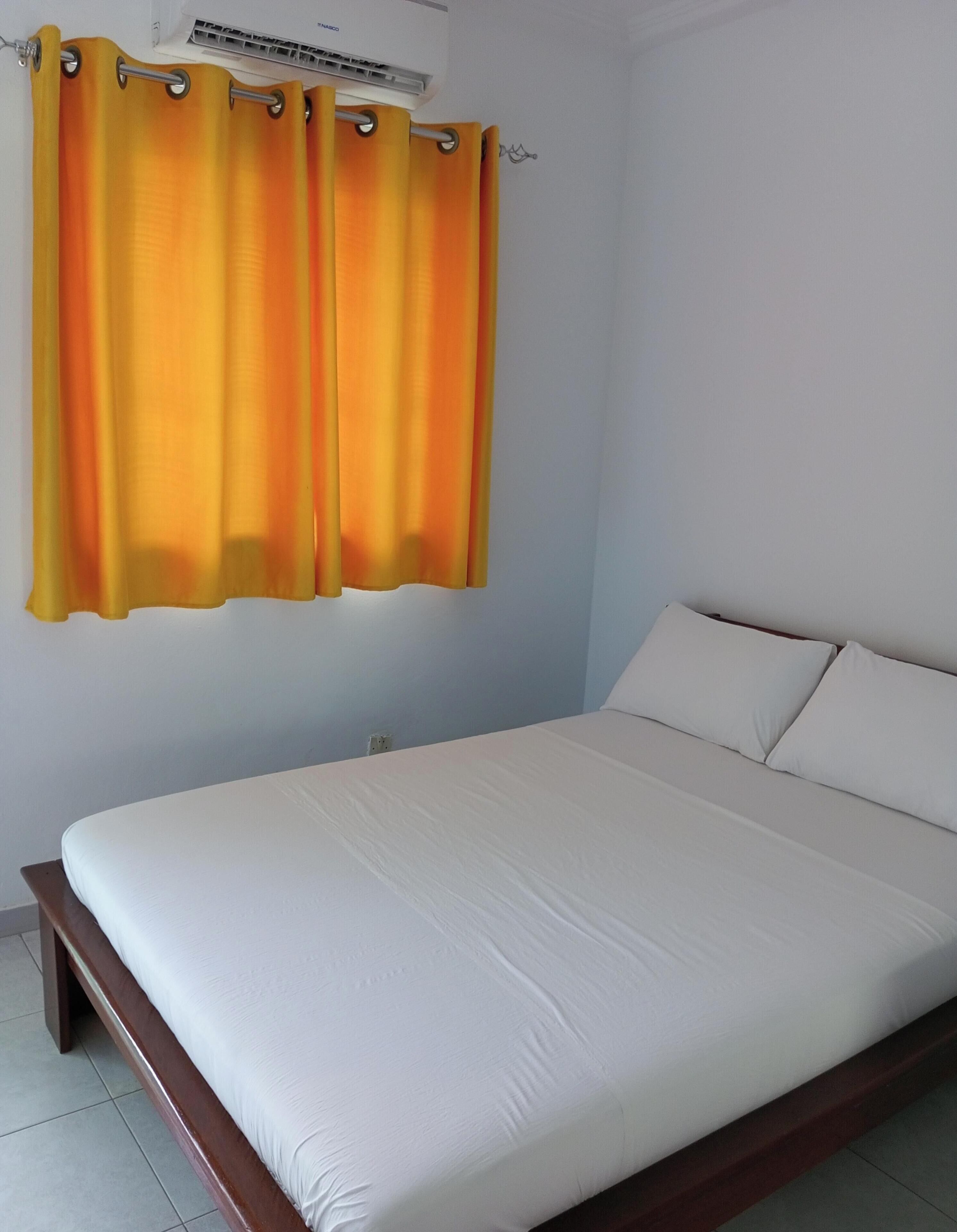 Een slaapkamer met een bed, witte gordijnen en een luchtkoeler aan de muur.