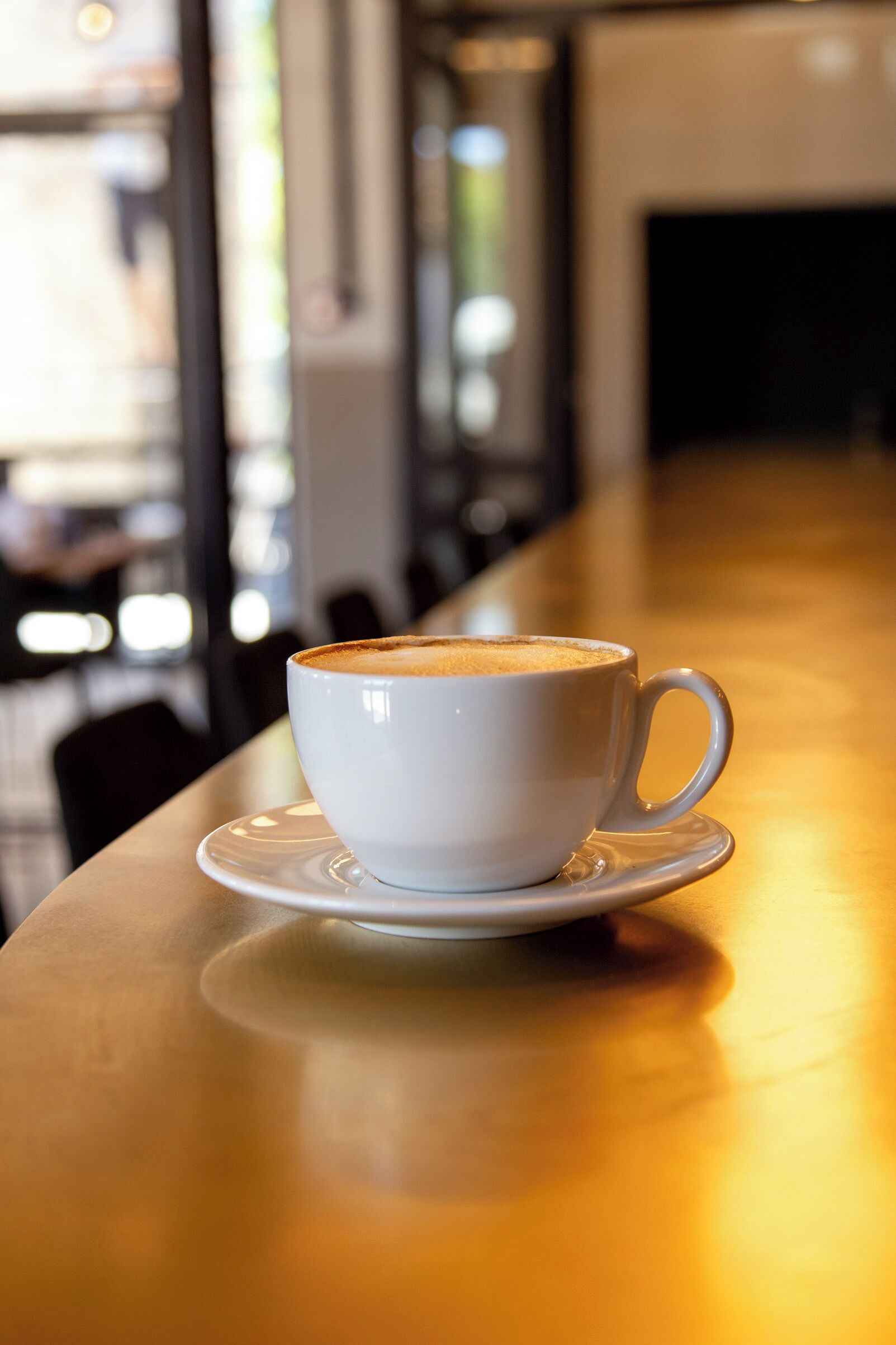 A white cup of coffee on a saucer placed on a wooden table in a cafe setting.