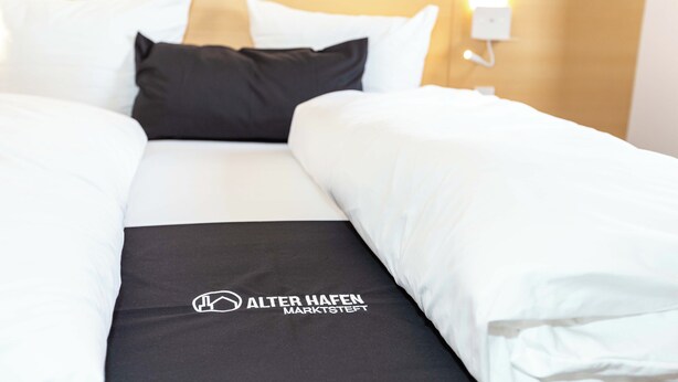 A hotel room with a bed, pillows, and a logo on the bedspread. - Alter Hafen Marktsteft (Marktsteft)